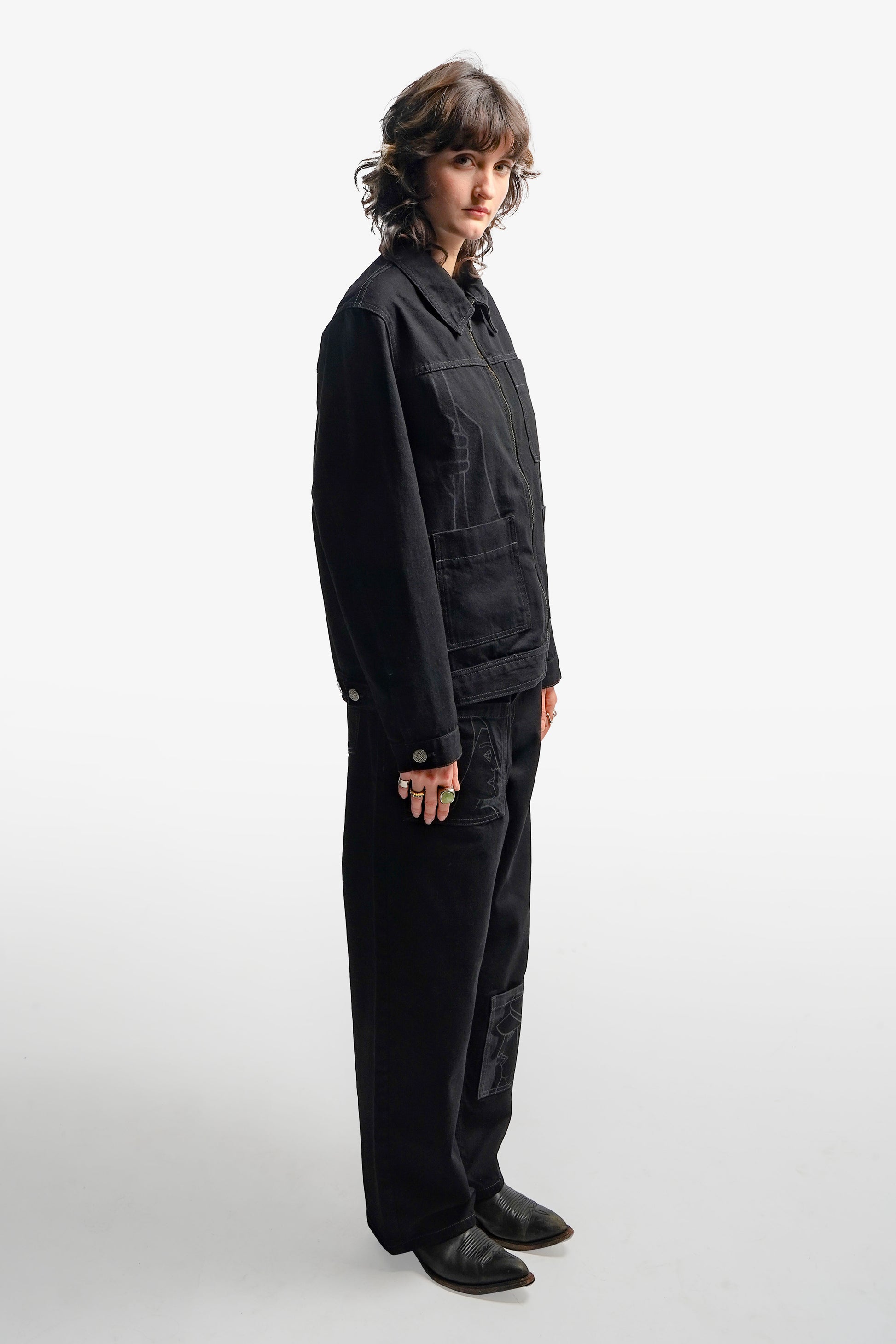 Veste en denim noir à l’inspiration workwear, revisitée avec une approche contemporaine. Son design structuré se distingue par trois poches carrées sur l’avant et des impressions au laser évoquant la fresque Avec Vous. Confectionnée en denim deep black rinse 100% coton, elle présente des surpiqûres en contraste gris, tandis qu’un jacron en cuir embossé noir apposé au dos signe son caractère unique.