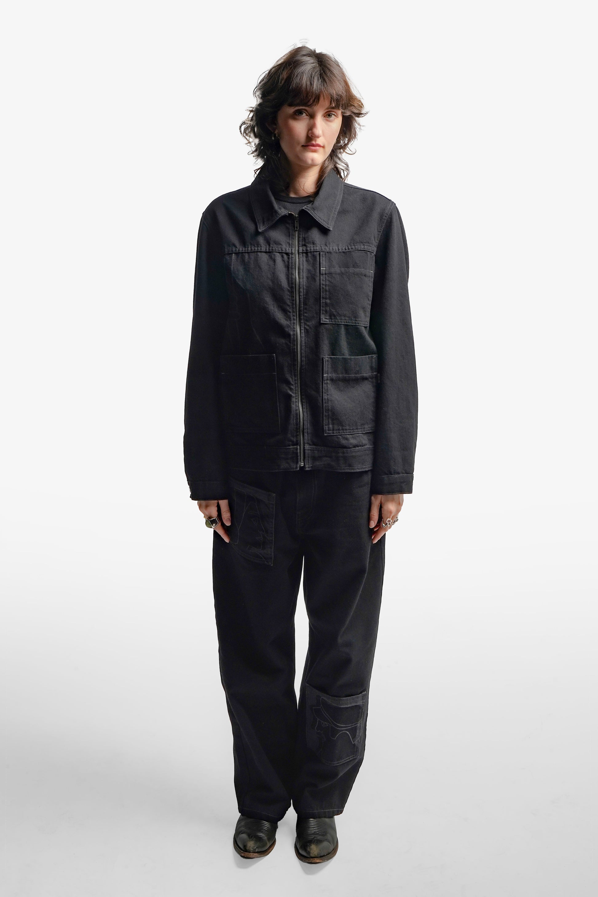 Veste en denim noir à l’inspiration workwear, revisitée avec une approche contemporaine. Son design structuré se distingue par trois poches carrées sur l’avant et des impressions au laser évoquant la fresque Avec Vous. Confectionnée en denim deep black rinse 100% coton, elle présente des surpiqûres en contraste gris, tandis qu’un jacron en cuir embossé noir apposé au dos signe son caractère unique.